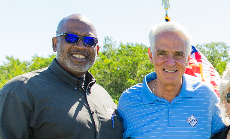 A photograph of Ken Welch and Charlie Crist smiling during an outdoor event. Ken Welch is on the left wearing sunglasses and a dark button-down shirt; Charlie Crist is on the right in a blue striped polo shirt.