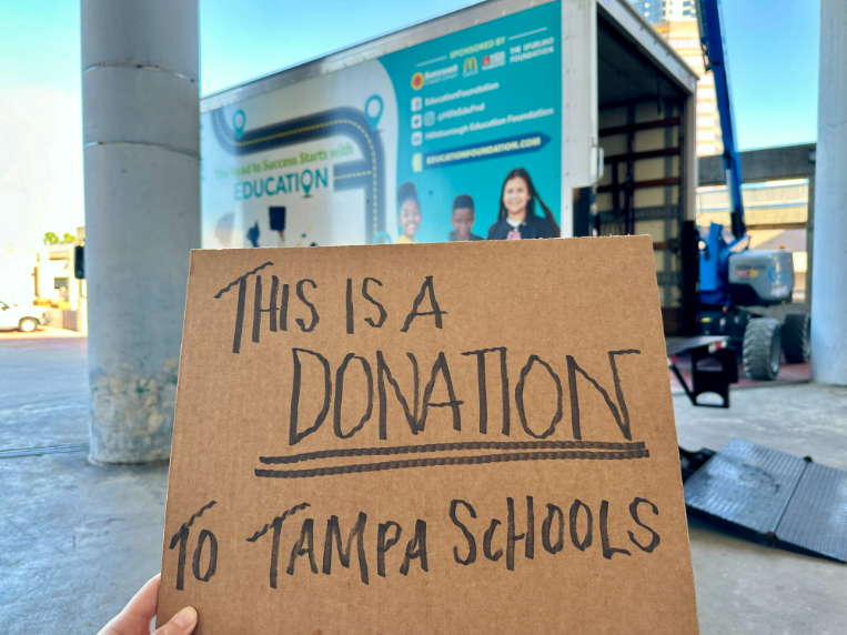 Sign on cardboard reads this is a donation to Tampa schools with a box truck in the background that depicts students on the side of it