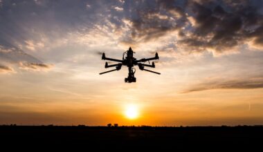 A large drone with multiple rotors flies in silhouette against an orange and yellow sunset sky, with scattered clouds and the sun low on the horizon.
