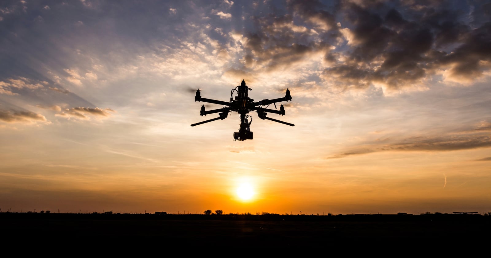 A large drone with multiple rotors flies in silhouette against an orange and yellow sunset sky, with scattered clouds and the sun low on the horizon.