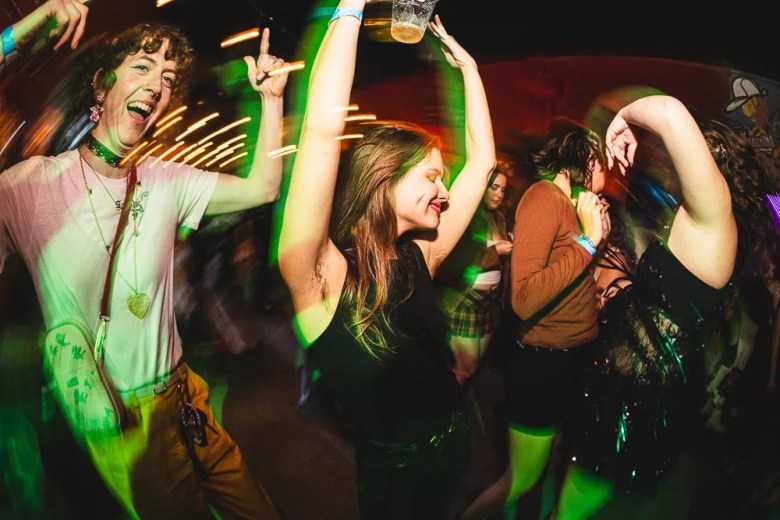 A vibrant, high-energy nightclub photograph of partygoers dancing at Dyke Night St. Pete on the patio of Crowbar. In the foreground on the left, an individual with short curly hair, wearing a light t-shirt, tan pants, a black choker, and a gold heart necklace, is smiling widely with one arm raised in the air. Next to them in the center, another individual with long brown hair, wearing a black sleeveless top, is dancing with both arms raised high above their head, holding a clear plastic cup in one hand and smiling with their eyes closed. The dark, lively venue is intensely illuminated by bright green light beams crossing the frame, along with horizontal orange light trails that capture the energetic movement of the dancers. Additional partygoers are visible dancing in the softly blurred background.