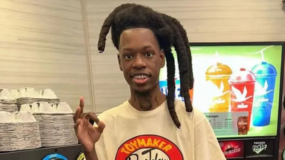 Julio Foolio with unique, tall dreadlocks and a beige shirt stands indoors, making a hand gesture, with colorful drink machines in the background.