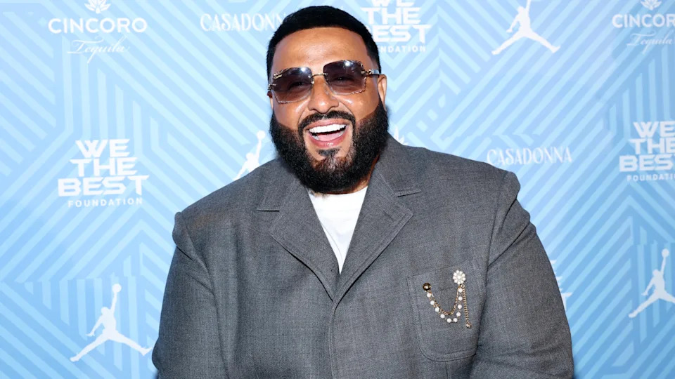 DJ Khaled smiling in a gray suit with sunglasses, against a blue patterned backdrop.