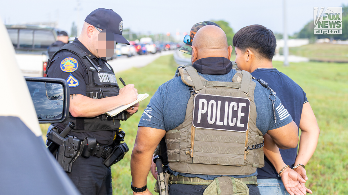A police officer writes on paperwork while standing beside a person in handcuffs during an immigration enforcement operation in Florida.
