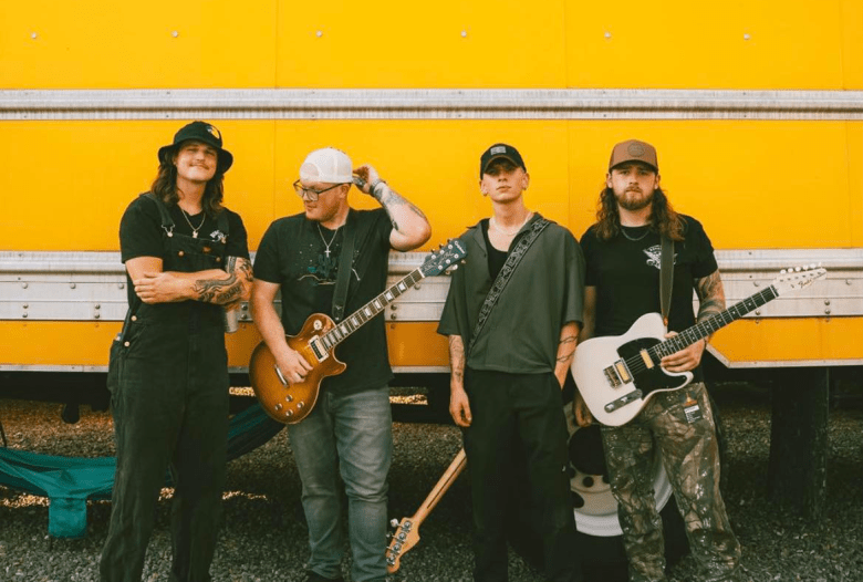 A bright, outdoor daytime portrait photograph of the four members of the band Fox n' Vead standing in a row in front of the brightly lit, solid yellow side of a large trailer. From left to right: the first member has long hair and a mustache, wearing a black bucket hat and black overalls over a t-shirt, smiling with their arms crossed. The second member is wearing a white backward baseball cap, glasses, and a black t-shirt, holding a sunburst-finish electric guitar with both hands. The third member is wearing a loose, olive-green short-sleeve button-down shirt and dark pants with their hands resting casually at their sides. The fourth member has a beard and is wearing a backward baseball cap, a black graphic t-shirt, and camouflage pants, holding a white electric guitar by the neck. The overall image has a warm, distinctly retro color grade.