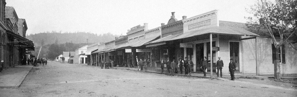 A dusty stretch of California Street in Jacksonville, where wooden storefronts, hitching posts, and a scattering of townspeople capture the rhythm of a late-1800s gold rush community settling into everyday life. Photo: Historic Jacksonville.