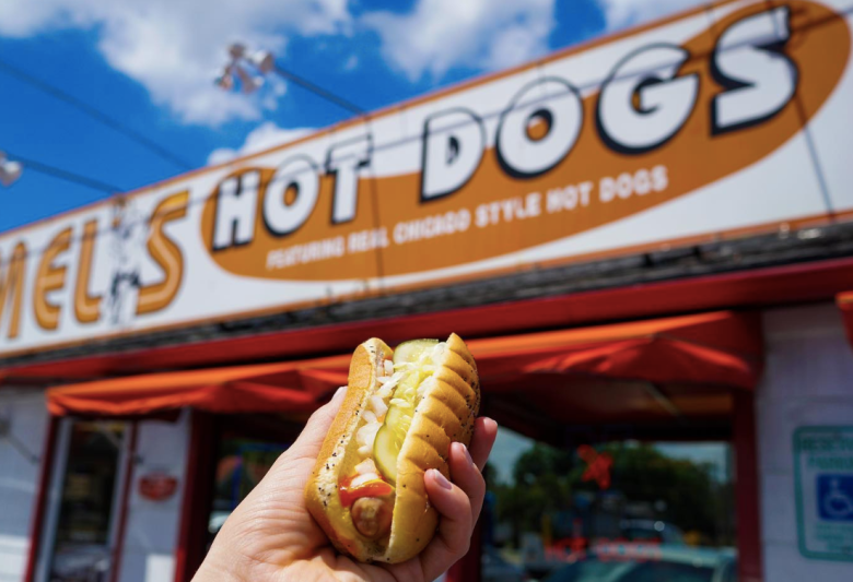 A hand holding a hot dog outside Mel's Hot Dogs