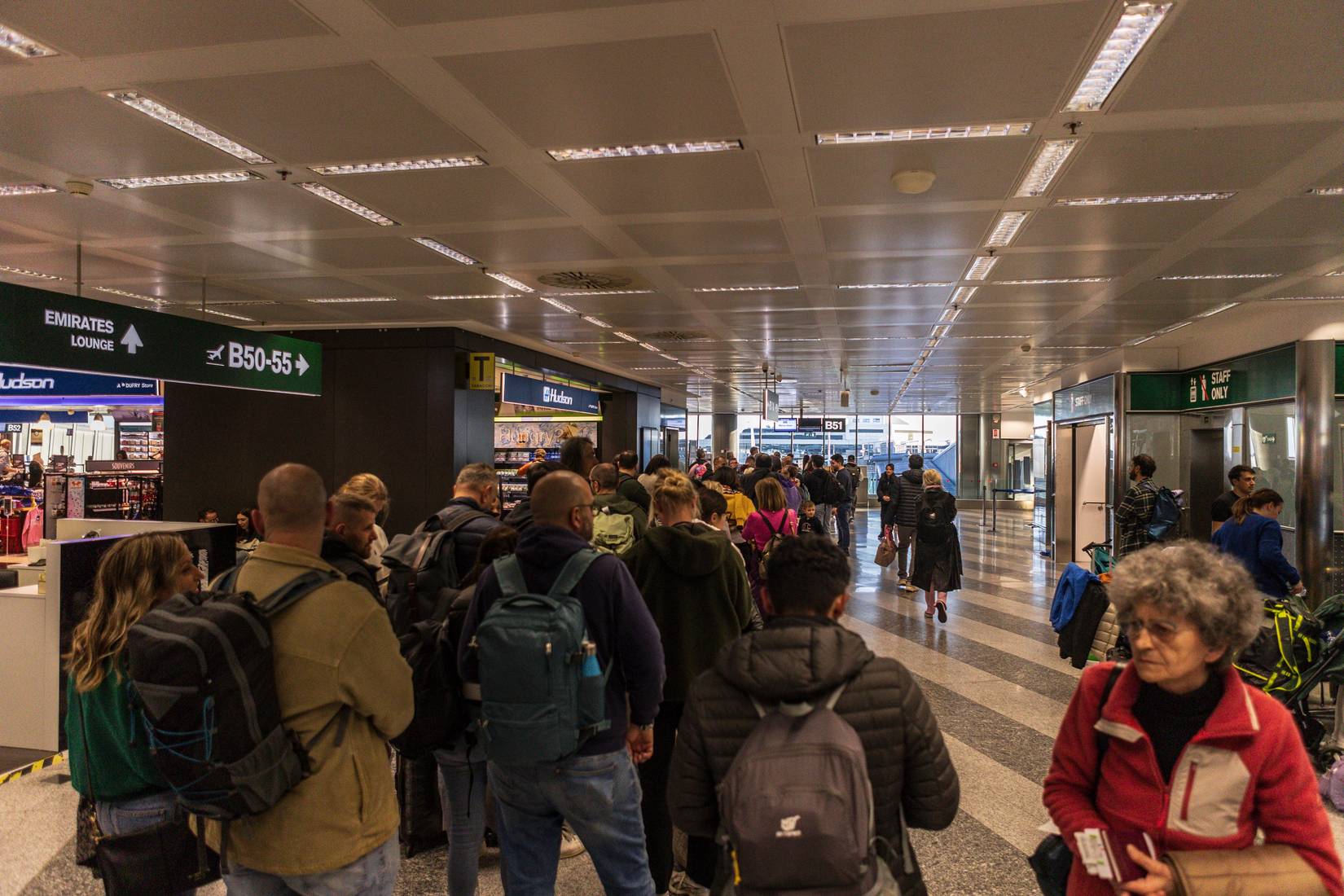 Tourists with backpacks and luggage waiting in line inside Milan Malpensa airport, surrounded by modern architecture and busy travelers