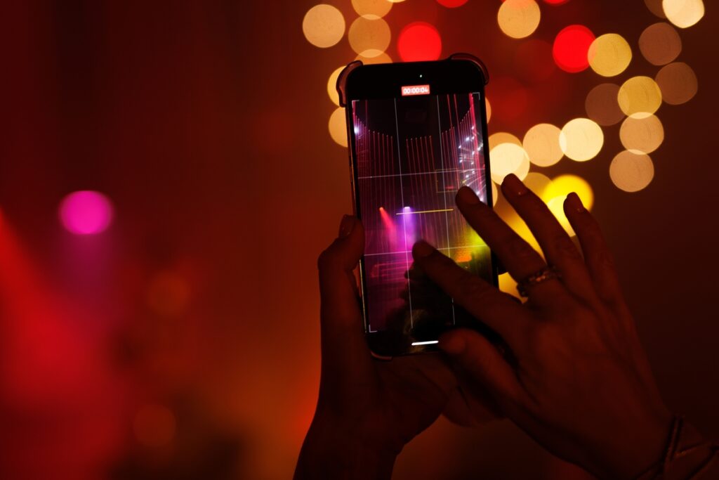 Hands holding a phone recording bright stage lighting in a dim room. Bokeh background and party mood in a nightlife setting.