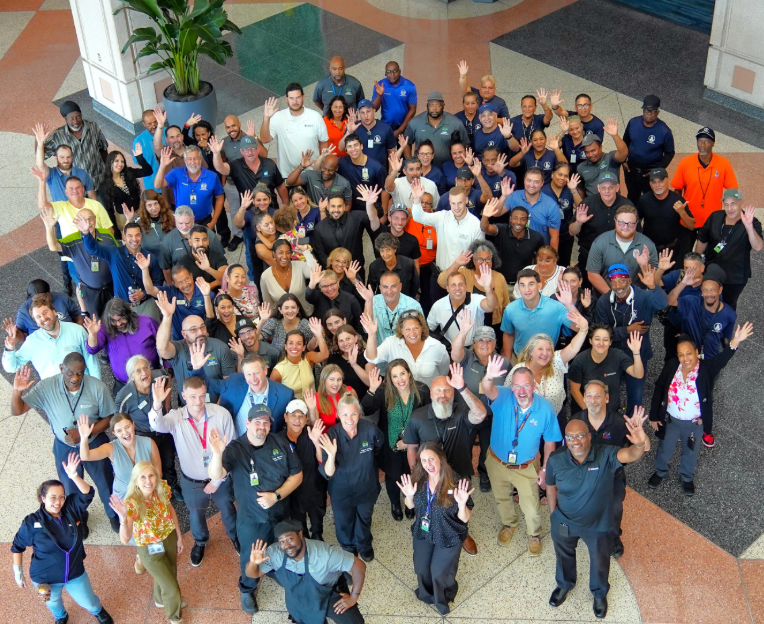 a group photo of professionals posing together indoors on a grand concrete staircase. the individuals are dressed in business casual attire and uniforms, smiling warmly and waving up at the camera