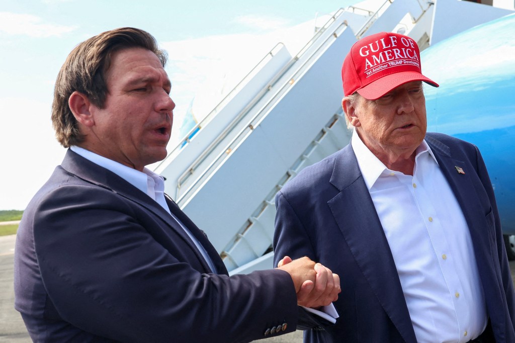Florida Governor Ron DeSantis holds hands with U.S. President Donald Trump, who is wearing a red hat that reads "GULF OF AMERICA Yet Another TRUMP Development."