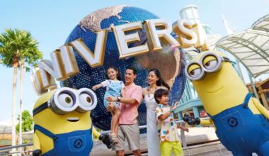 A smiling family of four, with two children held by parents, poses in front of the iconic Universal Studios globe. Two Minions character mascots stand on either side of the family. The backdrop includes palm trees and part of the new land at the theme park.