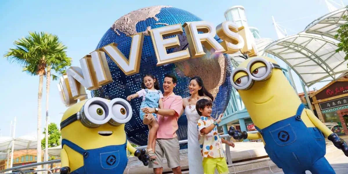 A smiling family of four, with two children held by parents, poses in front of the iconic Universal Studios globe. Two Minions character mascots stand on either side of the family. The backdrop includes palm trees and part of the new land at the theme park.