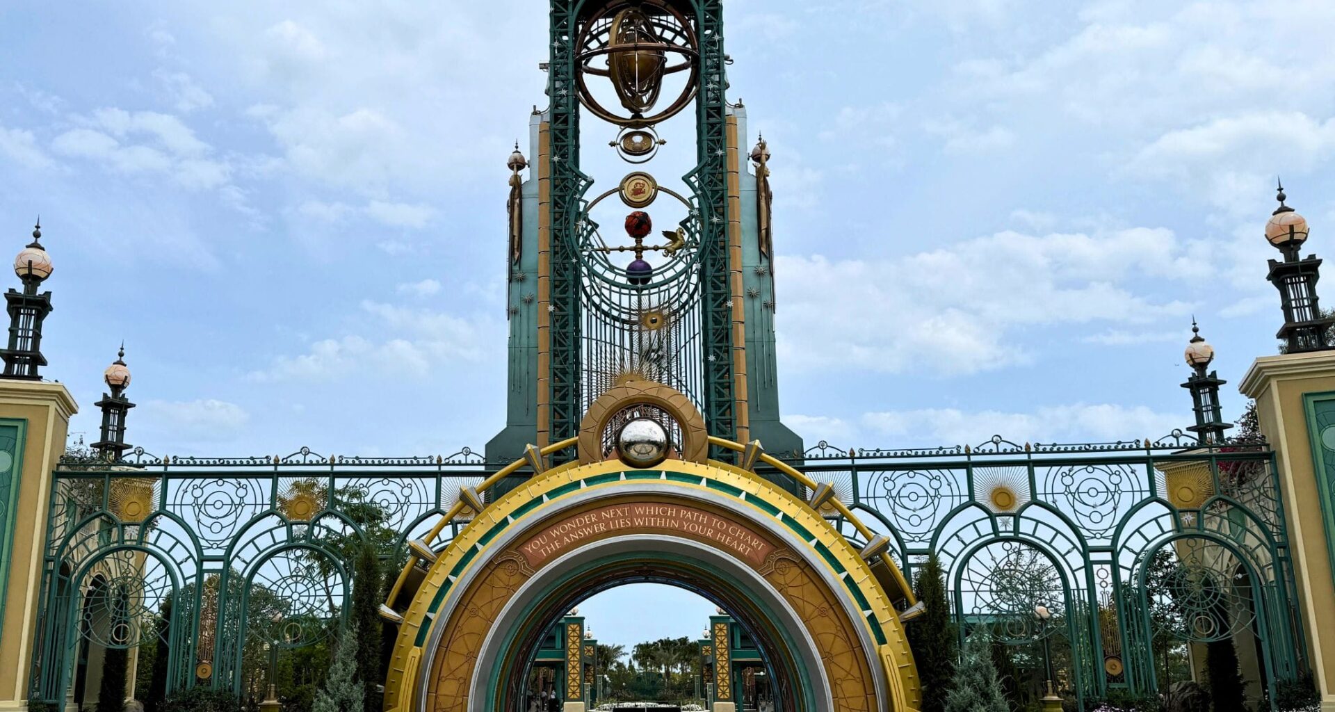 Ornate, fantasy-style entrance gate with tall arches, gold and green details, and decorative orbs under a cloudy sky.