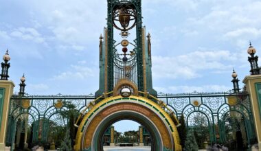 Ornate, fantasy-style entrance gate with tall arches, gold and green details, and decorative orbs under a cloudy sky.