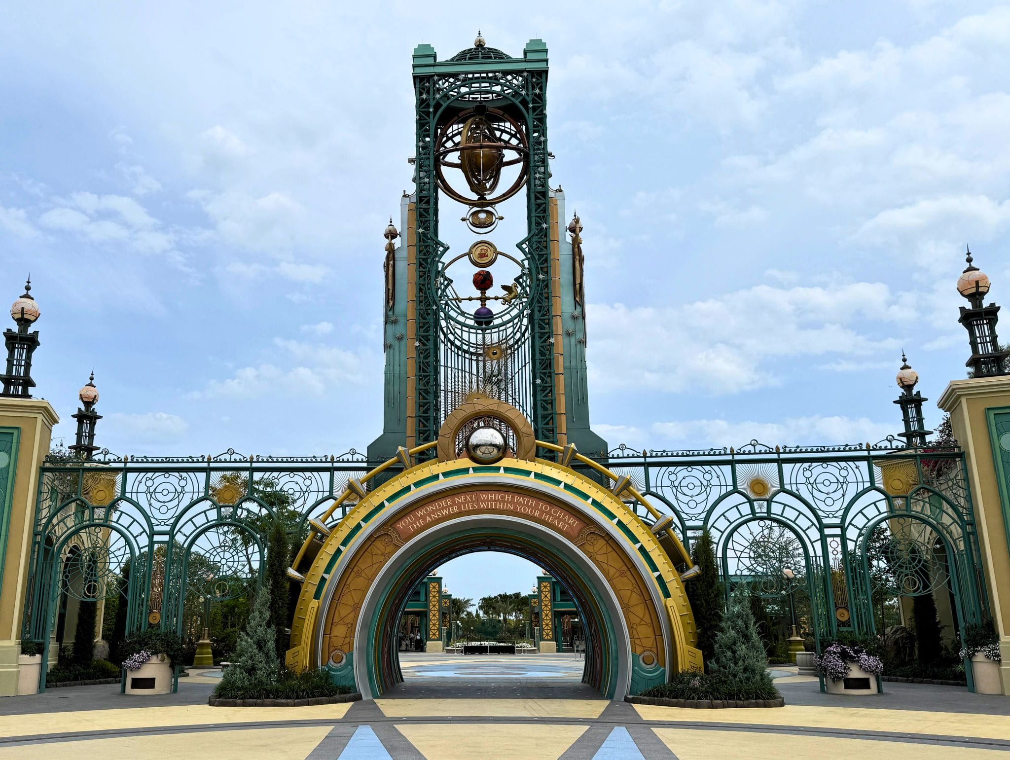 Ornate, fantasy-style entrance gate with tall arches, gold and green details, and decorative orbs under a cloudy sky.