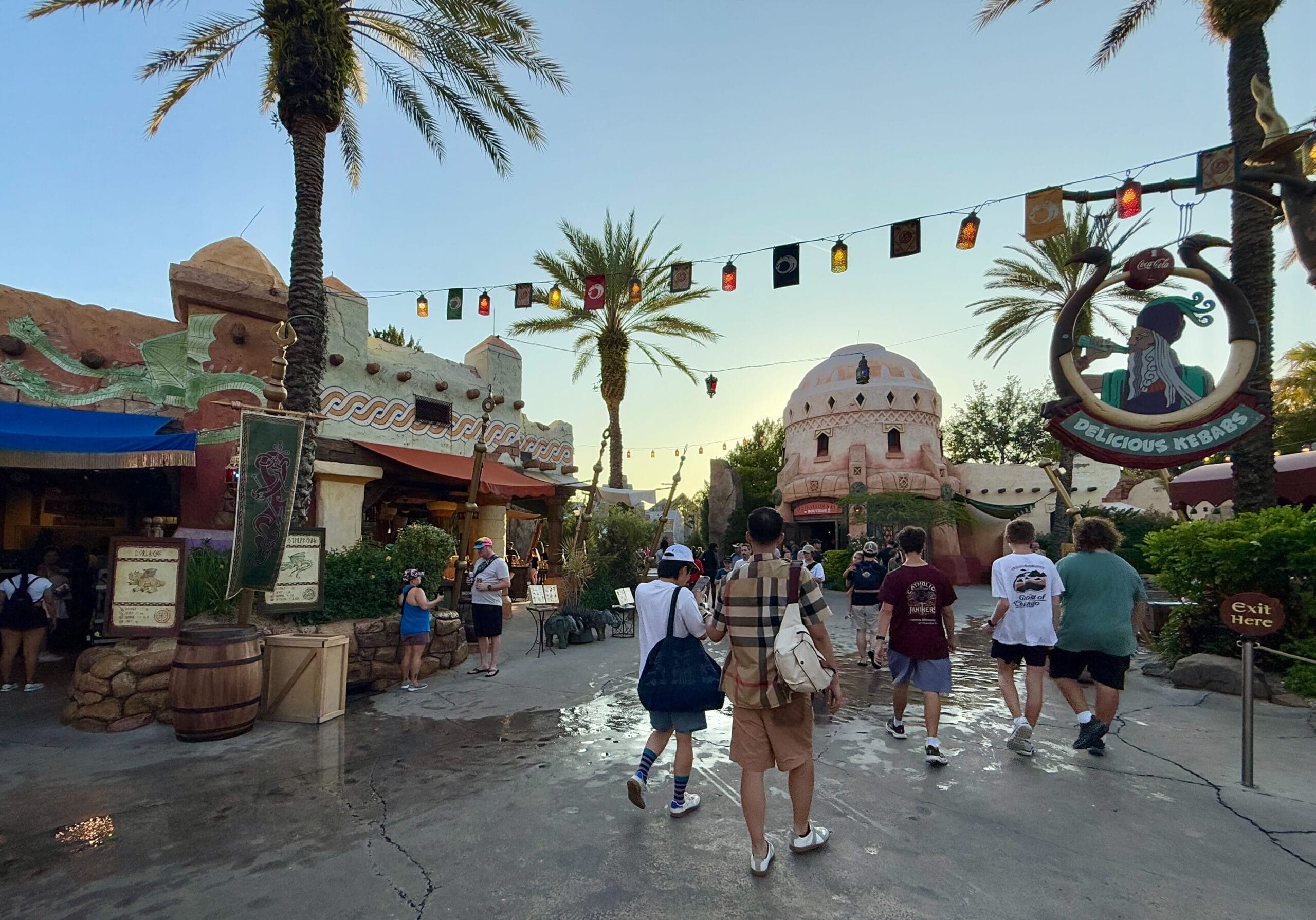 People stroll through The Lost Continent plaza with palm trees, colorful buildings, and lanterns at sunset.