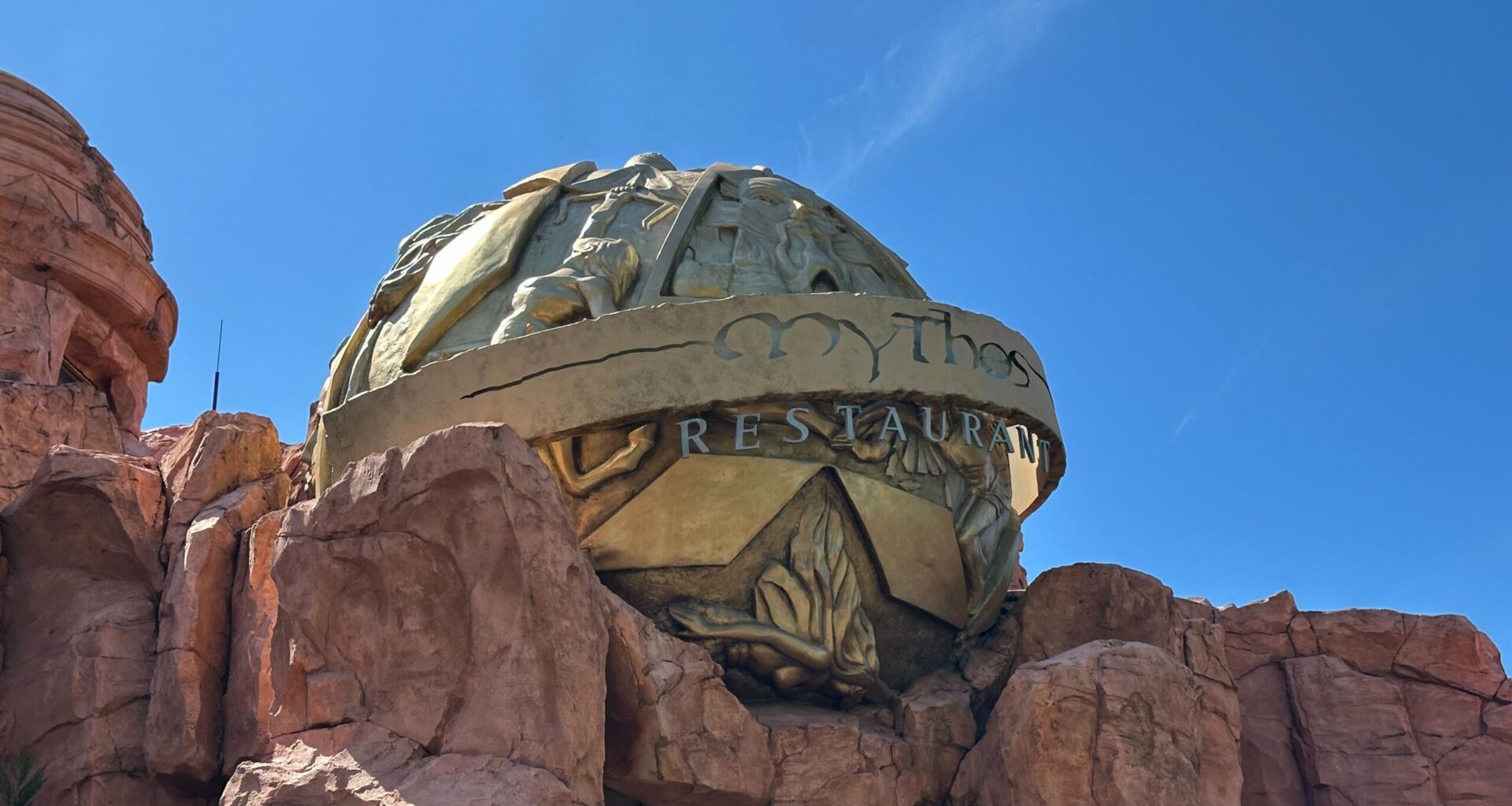 The exterior of Mythos Restaurant, adorned with a large, decorative golden dome and intricate reliefs, is nestled among rocky structures under a clear blue sky. It captures the holiday spirit at Universal Islands of Adventure, adding to its festive allure during Christmas.