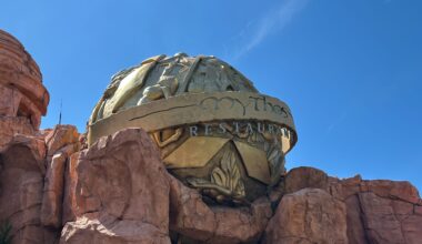 The exterior of Mythos Restaurant, adorned with a large, decorative golden dome and intricate reliefs, is nestled among rocky structures under a clear blue sky. It captures the holiday spirit at Universal Islands of Adventure, adding to its festive allure during Christmas.