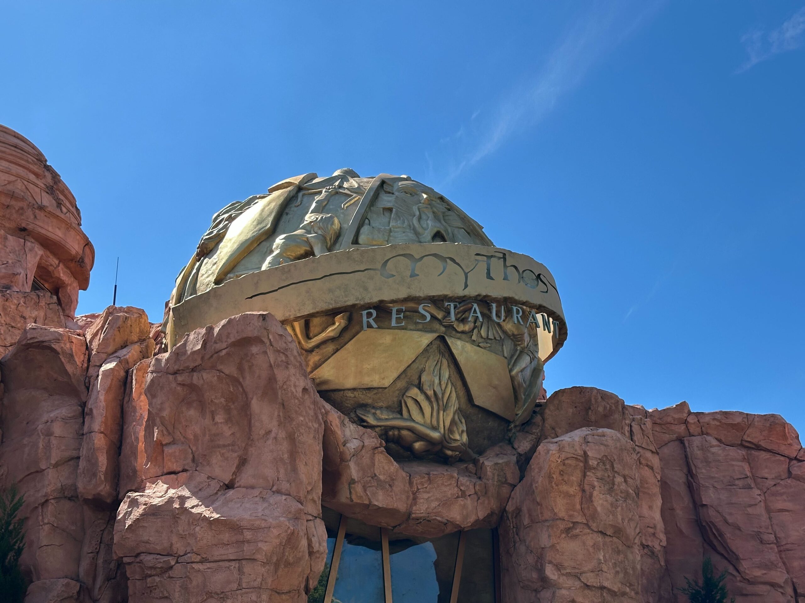The exterior of Mythos Restaurant, adorned with a large, decorative golden dome and intricate reliefs, is nestled among rocky structures under a clear blue sky. It captures the holiday spirit at Universal Islands of Adventure, adding to its festive allure during Christmas.
