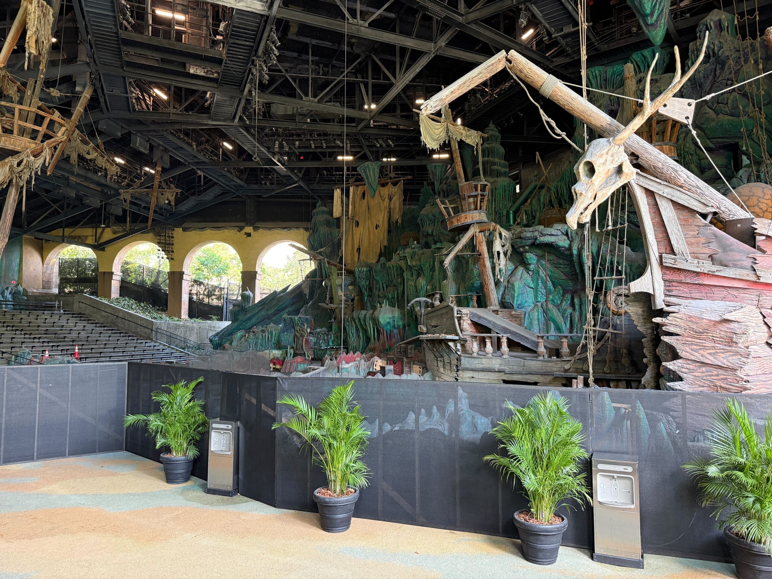 Large stage set with a pirate ship, jungle scenery, empty seats, and potted plants in front of a barrier.