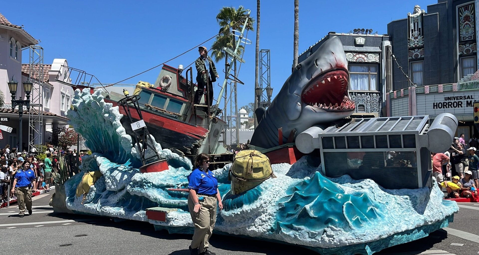 Parade float at the 2025 Universal Mega Movie Parade featuring a large shark, a red boat with crashing waves, and a person standing on the deck. Spectators and staff from Universal Orlando Resort are visible nearby under a clear blue sky.