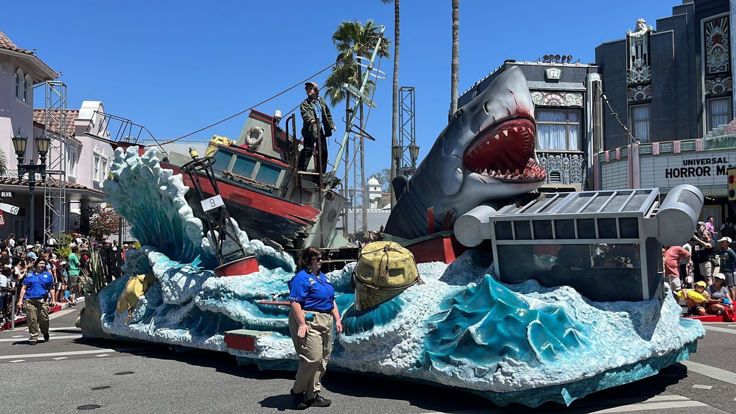 Parade float at the 2025 Universal Mega Movie Parade featuring a large shark, a red boat with crashing waves, and a person standing on the deck. Spectators and staff from Universal Orlando Resort are visible nearby under a clear blue sky.