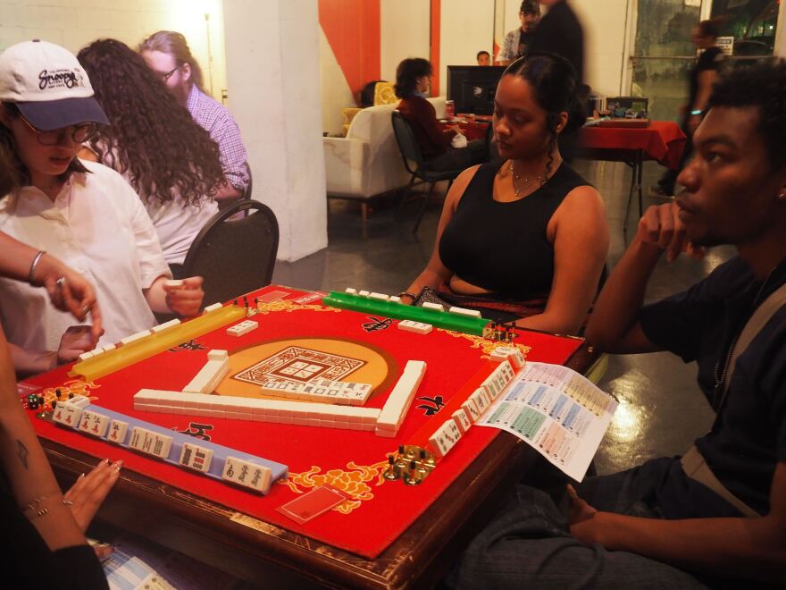 a group of players sit at a mahjong table, listening to instructions on how to play