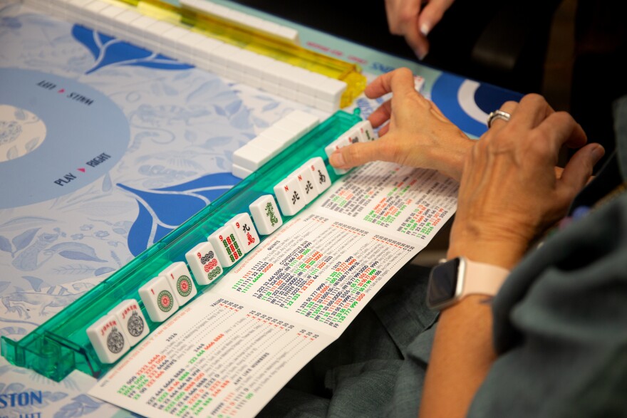 close up of hands grabbing a set of tiles on the mahjong table with the annual mahjong card placed on the table