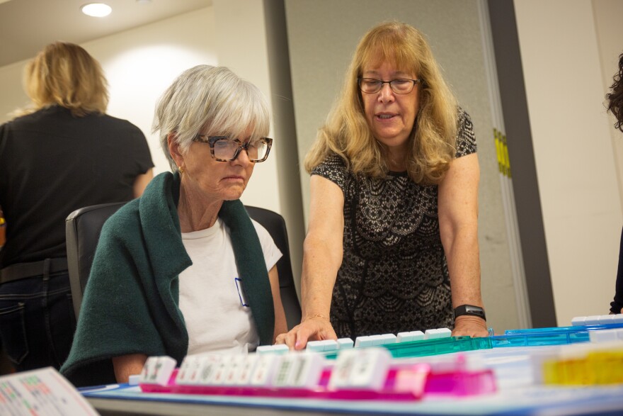 a woman stands beside a mahjong player at the table, pointing to a tile, during a mahjong lesson