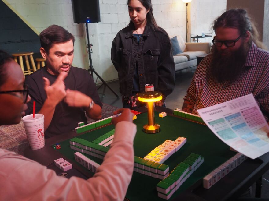 players sit around a mahjong table while an instructor stands beside them