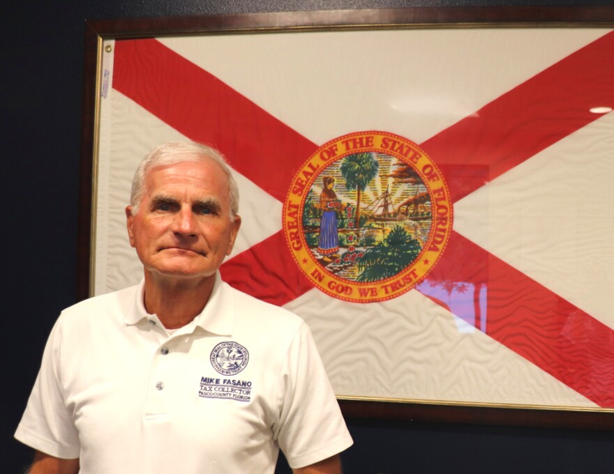 A man in a white polo shirt stands in front of a framed Florida flag. His shirt bears his name, "Mike Fasano, Tax Collector, Pasco County, Florida."