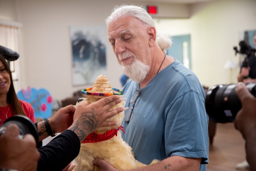 An older man with white hair in a ponytail and wearing a blue t-shirt holds a small, yellow robotic dog with a red bandana as a woman attaches a sombrero to its head.