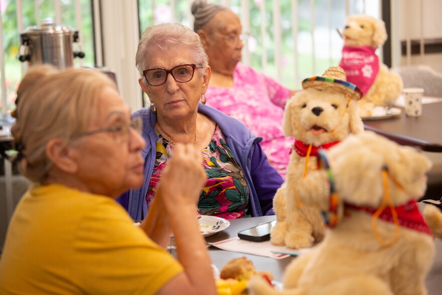 A woman with short gray hair and wearing glasses and a floral shirt with a purple hoodie over sits at a table with her little yellow dog with a red bandana and sombrero. Other woman, out of focus, sit around her with their dogs.