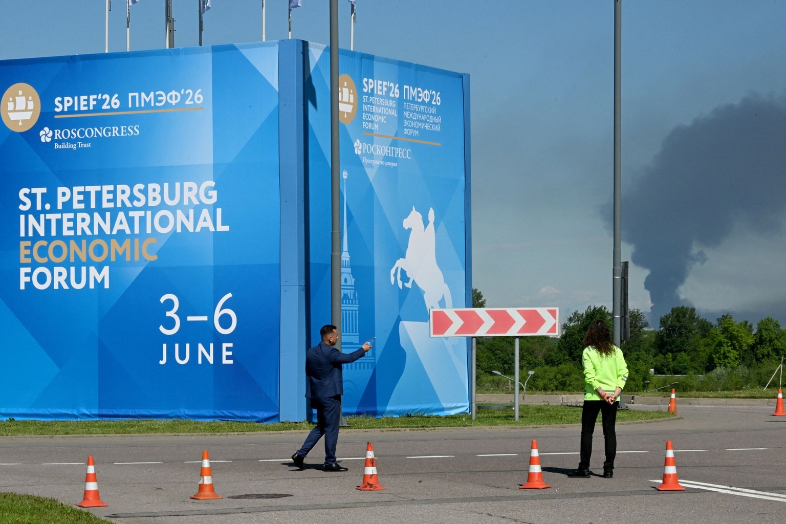 People stand in front of banners for the St. Petersburg International Economic Forum as black smoke rises in the distance in Saint Petersburg, Russia, on June 3, 2026.