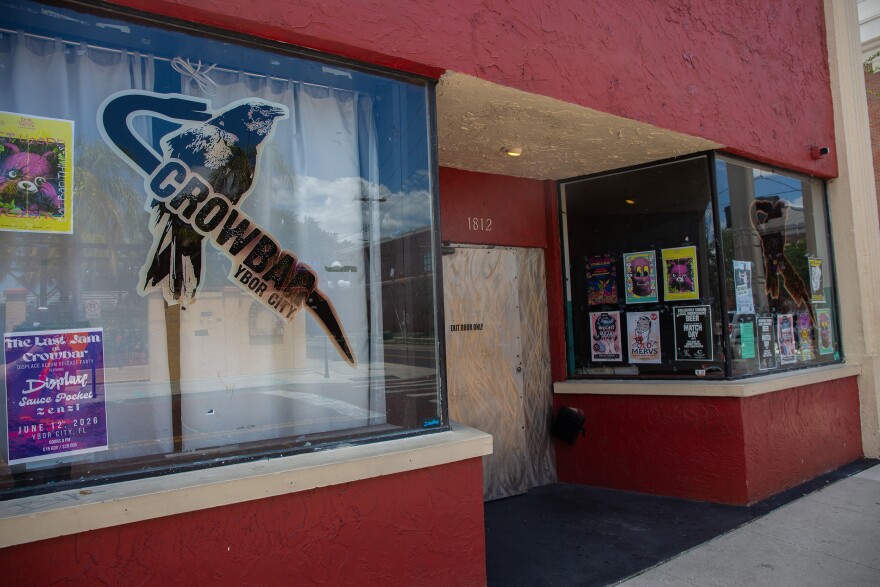 A side angle of the front entrance to a music venue. It has large glass panels with posters plastered to them. The building is dark red. A logo with a crow and a crowbar is on the front of it.
