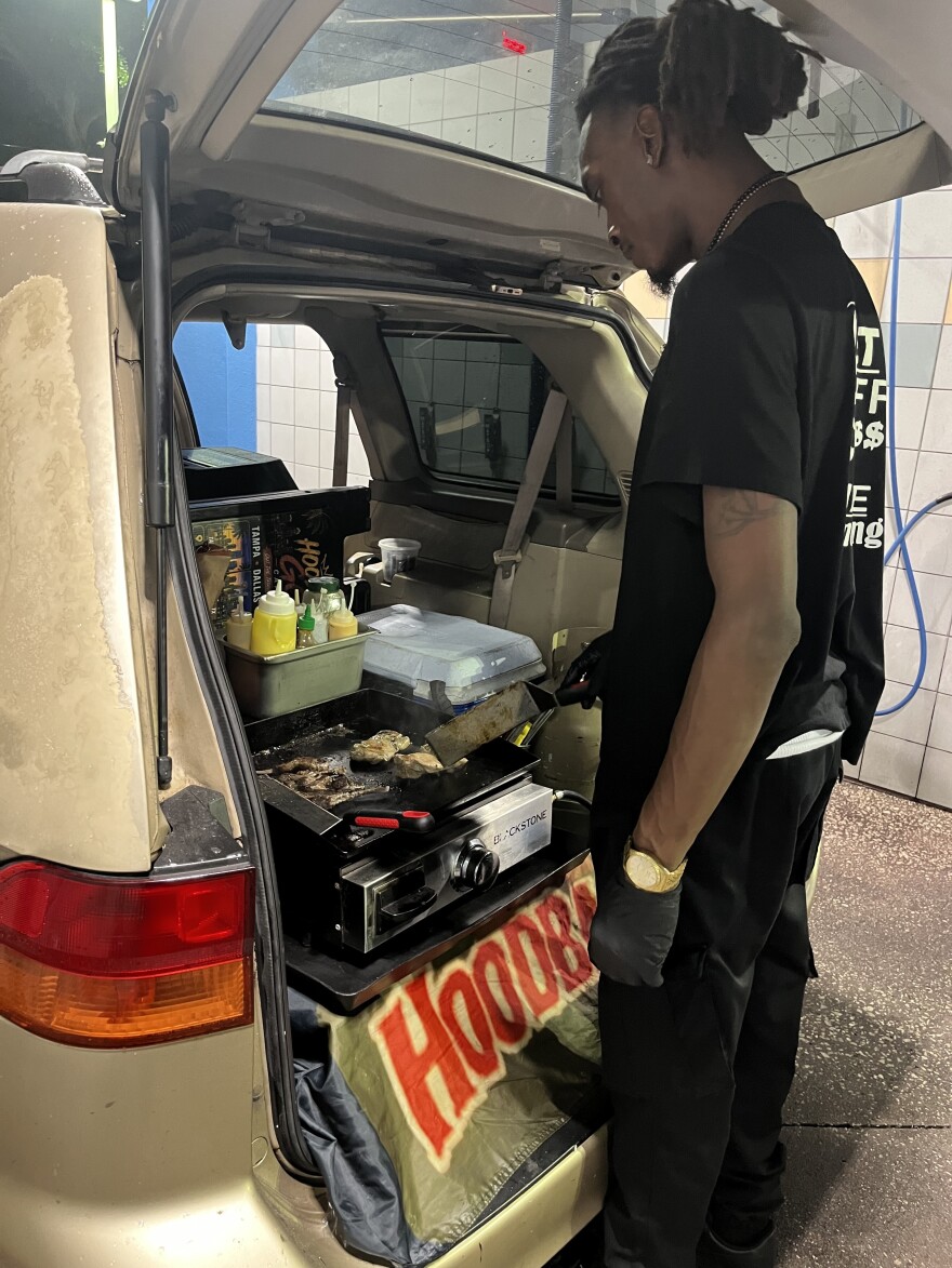 A Black man wearing a black shirt and gloves holds a plastic food box with shrimp and vegetables. You can see a car trunk with a grill stove installed, and a metal container containing various sauce bottles.