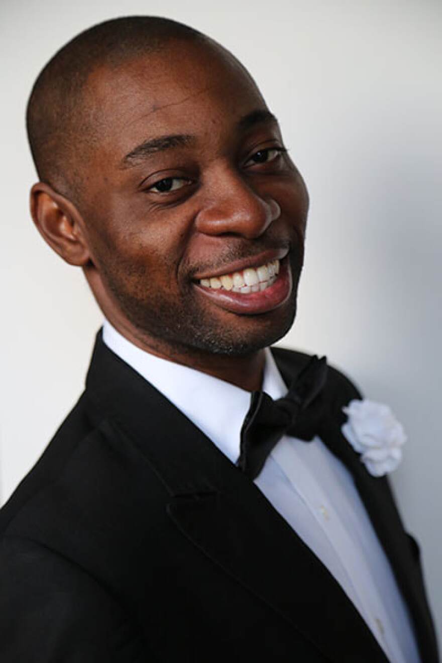 A young Black man dressed in black tie with a white boutonniere smiles at the camera.