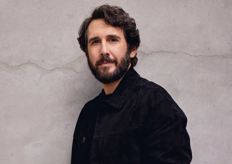 Josh Groban stands against a light gray concrete wall, facing the camera with a relaxed, thoughtful expression. His dark wavy hair and neatly trimmed beard frame his face, and he wears a dark suede jacket over a black shirt. Soft, even lighting and the minimalist background create a warm, understated portrait.