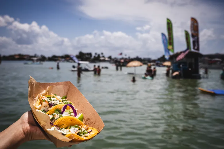 A paper basket of colorful fish tacos held up with Tiki's Taco Boat in the background