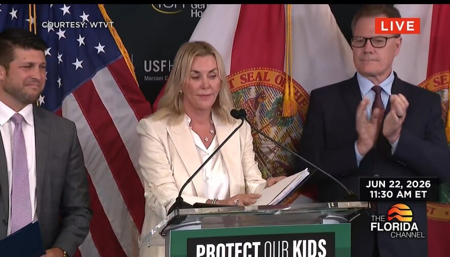 A blonde woman in a cream colored jacket speaks at a mic