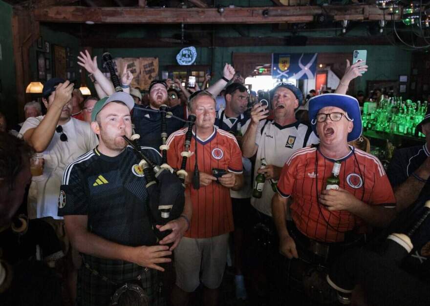 A group of men in soccer shirts sing inside a bar, one plays the bagpipe.