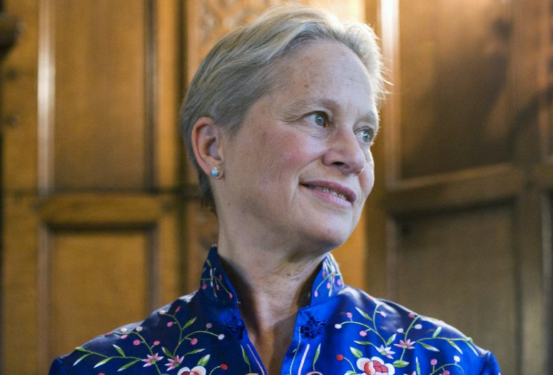 A portrait of classical pianist Rebecca Penneys looking slightly upwards and to the right with a soft smile. The musician has short, greyish-blonde hair and is wearing a striking, royal blue high-collared shirt featuring intricate pink, red, and white floral embroidery. The background consists of elegant, warm-toned wood paneling bathed in soft lighting.