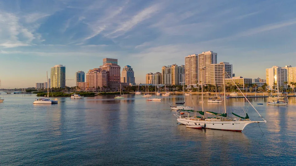 Sailboats anchored in a calm harbor with a city skyline in the background at sunset.