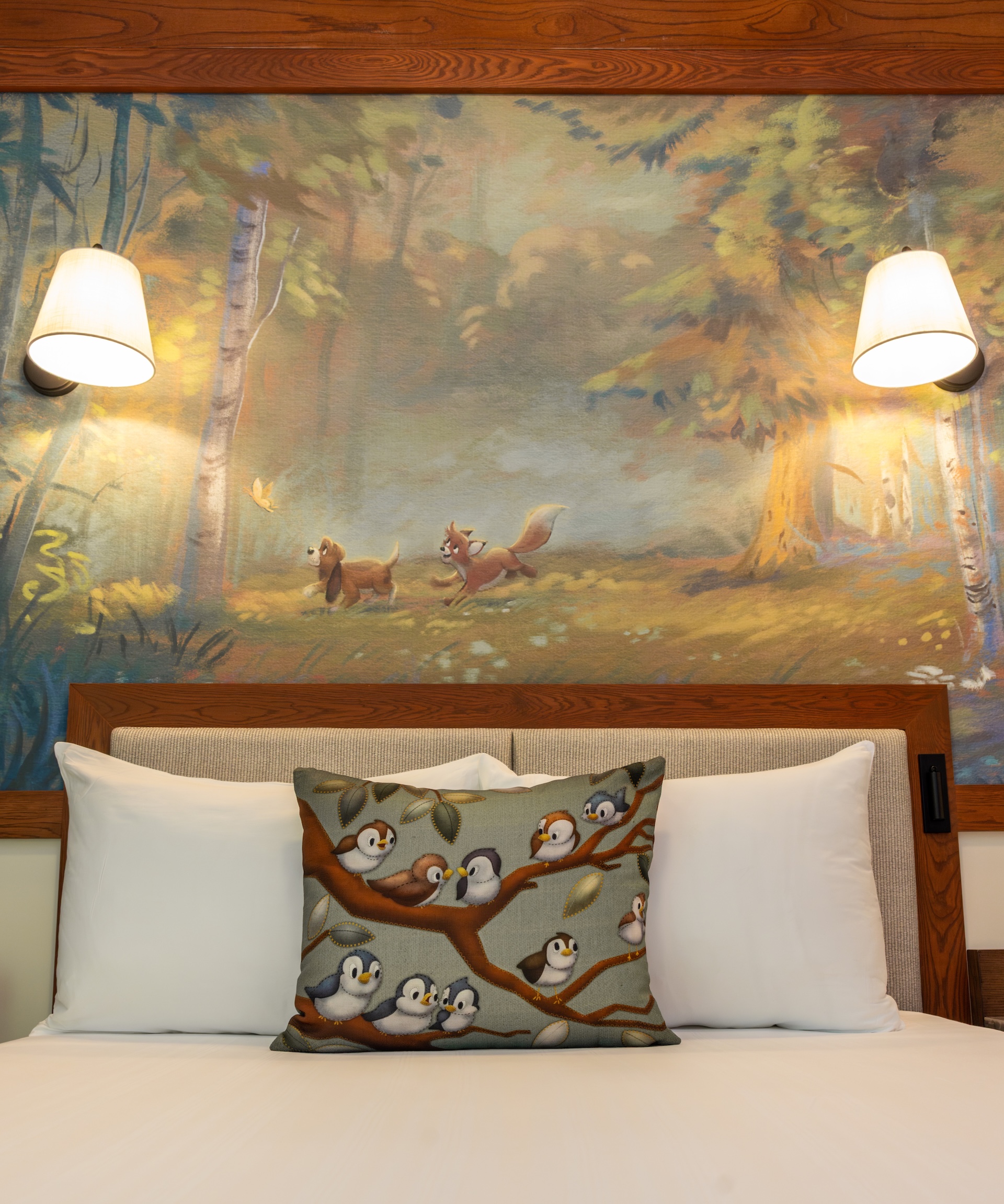 Bed with White Pillows and a Decorative Penguin print Cushion on a Branch Set Against a Forest Mural and Warm Wall Lamps