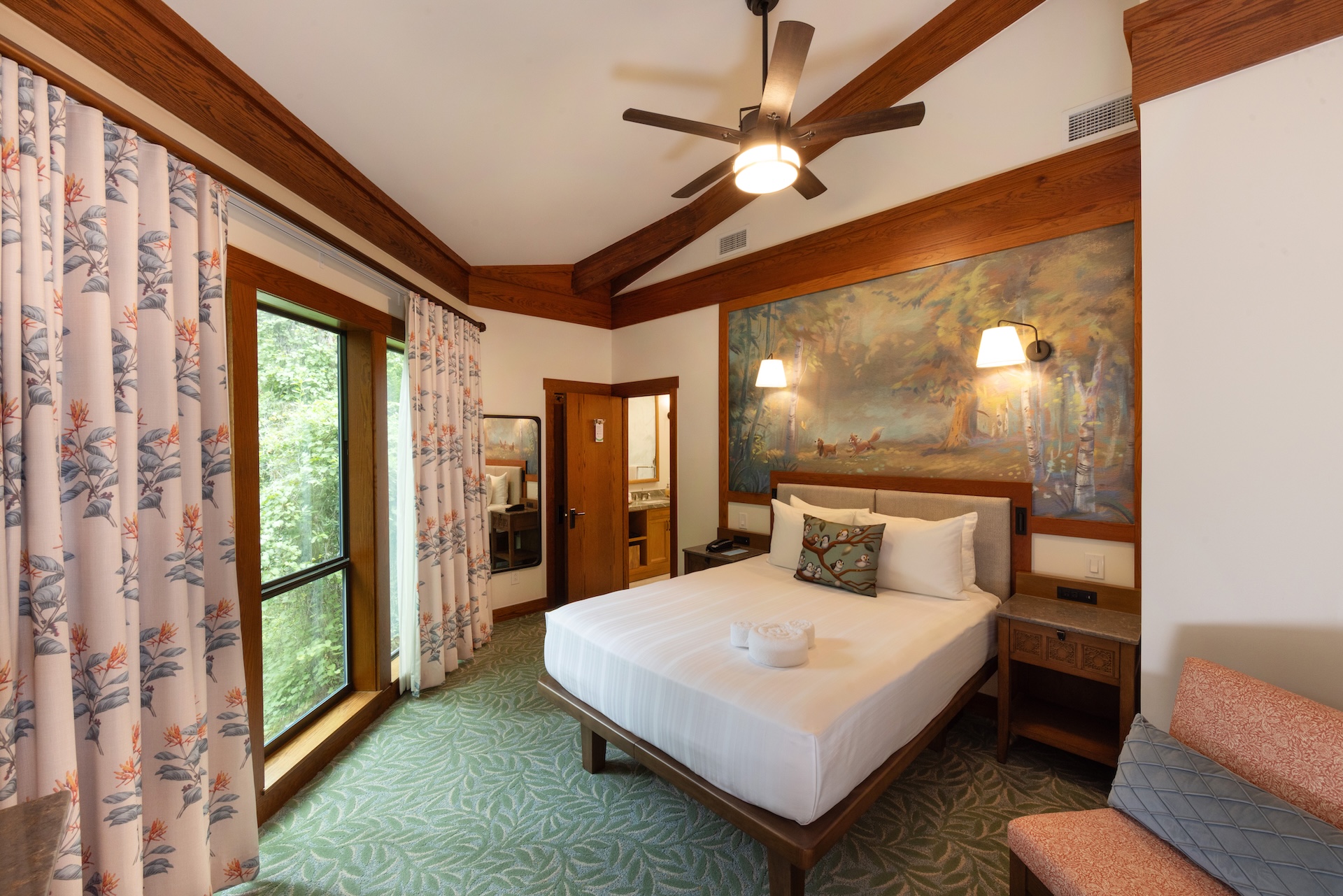 Hotel Room Featuring a King Bed with White Linens Wooden Headboard and a Large Forest Mural Above the Bed Window with Floral Curtains to the Left and Ceiling Fan Overhead
