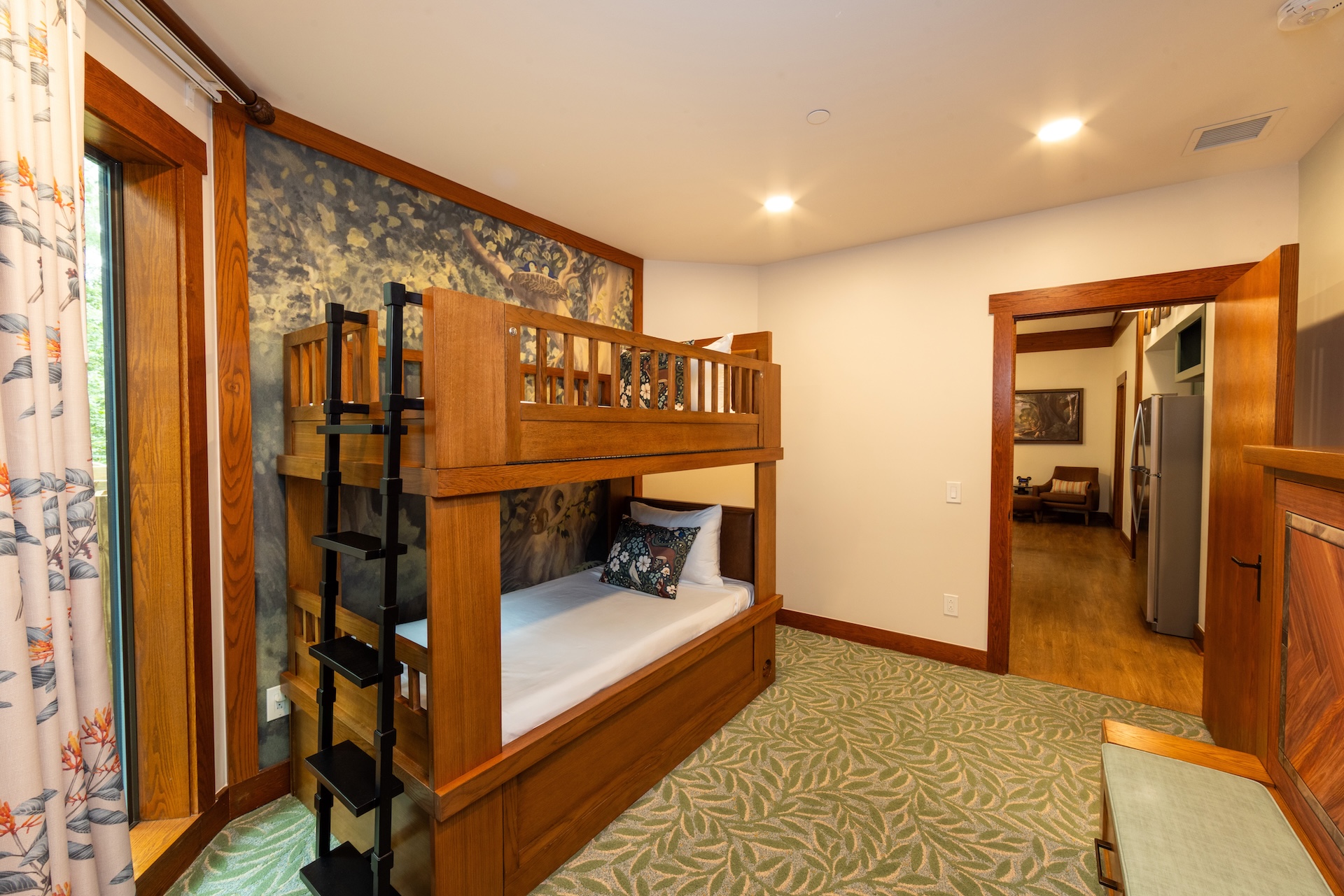 Wooden Bunk Bed with Metal Ladder in a Warmly Lit Room Green Patterned Carpet and a Doorway to a Living Areakitchen Beyond informative