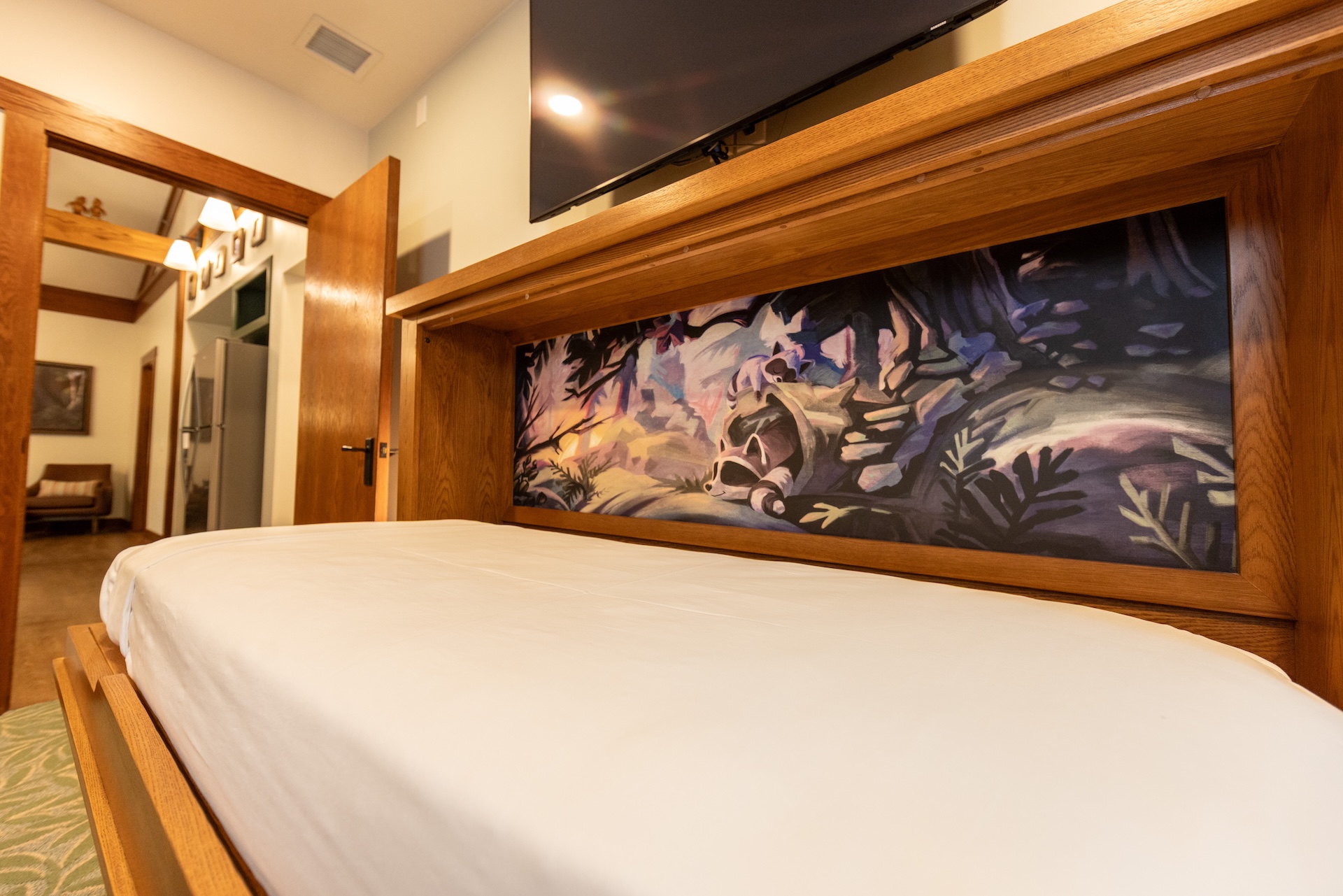 Low angle View of a Large Bed with a Wooden Headboard and a Colorful Mural Panel Behind It Tv Mounted Above Doorway and Hallway to the Left