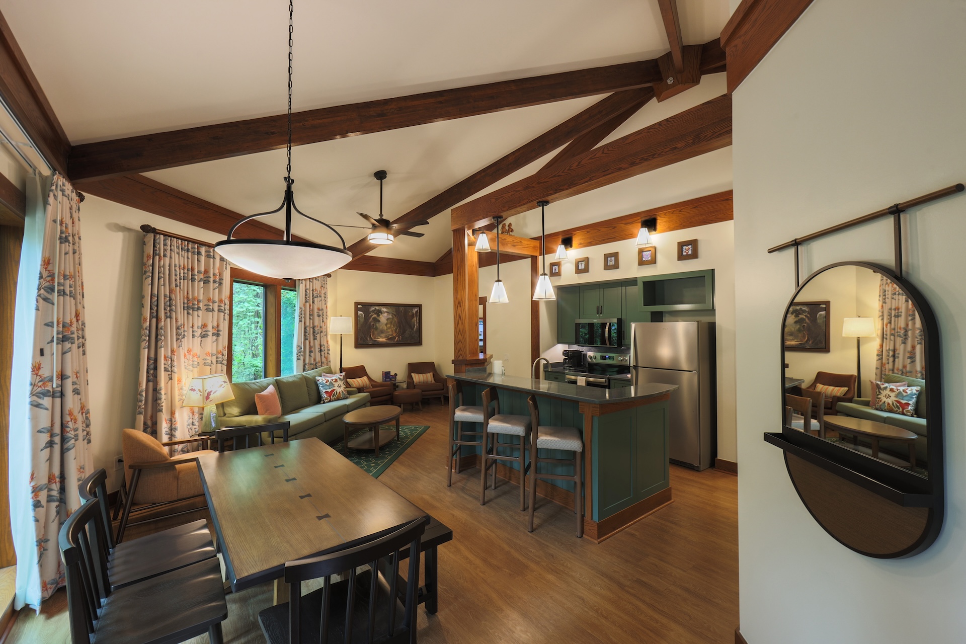 Open plan Living Area with Exposed Wooden Beams a Green Kitchen Island and a Dining Table Large Windows with Floral Curtains on the Left and a Round Wall Mirror on the Right