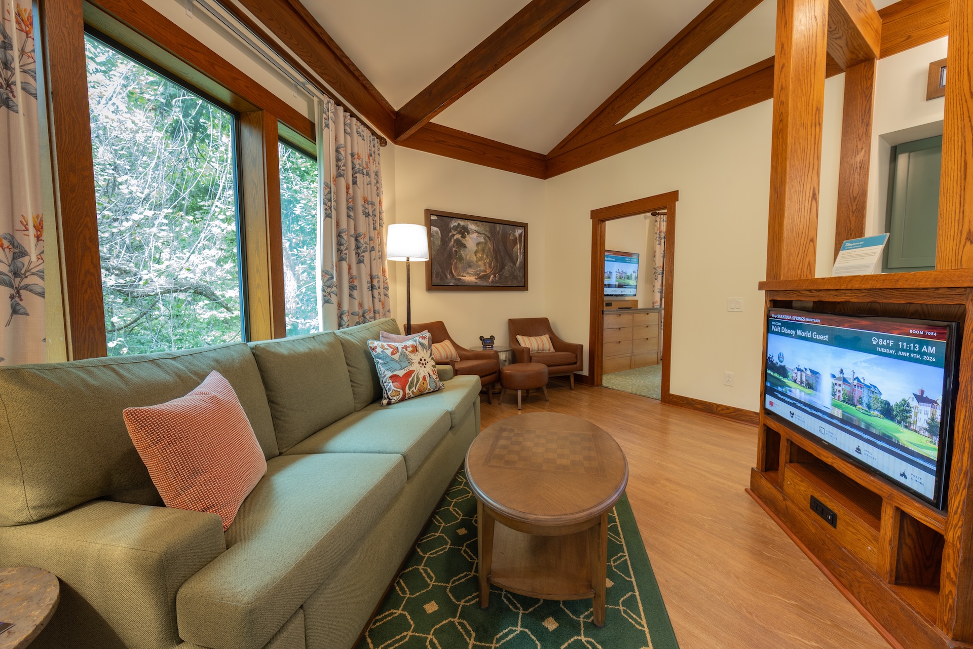 Living Room with a Green Sectional Sofa Pink Pillows and a Round Wooden Coffee Table Near Large Windows Showing Green Trees Outside